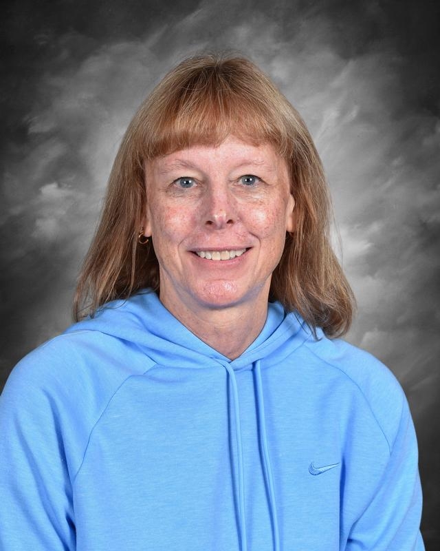 A smiling woman with light brown hair wearing a blue hoodie, set against a gray background.