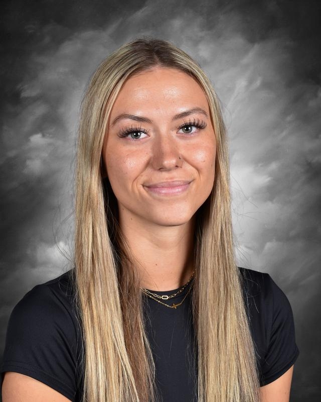 A young woman with long blonde hair smiles, wearing a black shirt, against a dark, cloudy background.