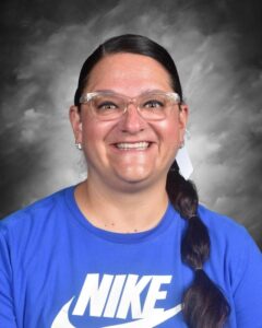 A smiling woman with glasses and long hair in a ponytail wears a blue Nike shirt against a gray background.
