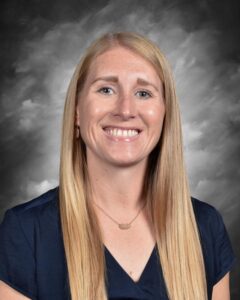 Smiling woman with long blonde hair, wearing a navy blue shirt, against a gray background.