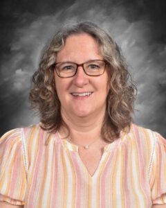 A smiling woman with curly hair, wearing glasses and a striped shirt, poses against a gray background.