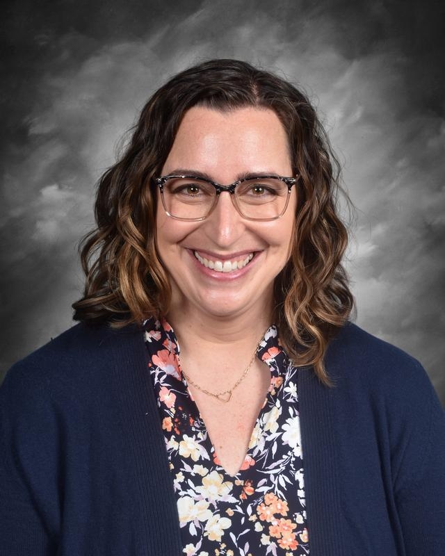 Smiling woman with curly hair, glasses, and a floral blouse, wearing a navy cardigan against a gray background.