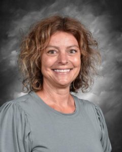Smiling woman with curly hair wearing a gray shirt, against a dark, cloud-like background.