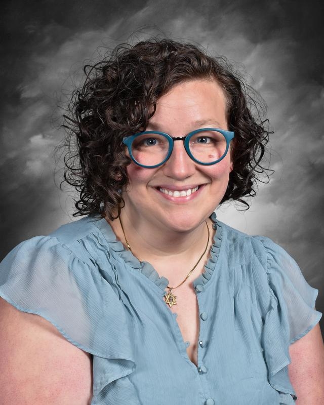 A smiling woman with curly hair wears blue glasses and a light blue blouse, against a gray background.
