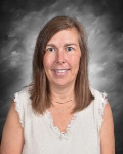 A woman with medium-length brown hair smiles at the camera, wearing a light-colored sleeveless top against a gray backdrop.