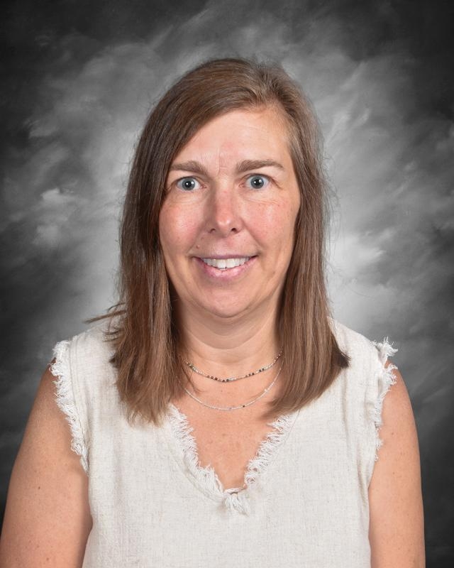 A woman with medium-length brown hair smiles at the camera, wearing a light-colored sleeveless top against a gray backdrop.