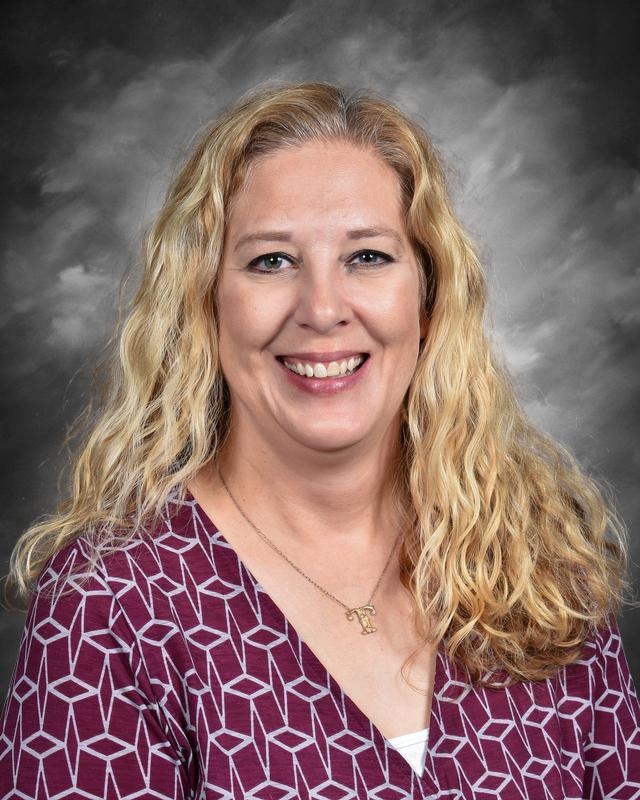 Smiling woman with long, curly blonde hair wearing a patterned top, against a gray background.