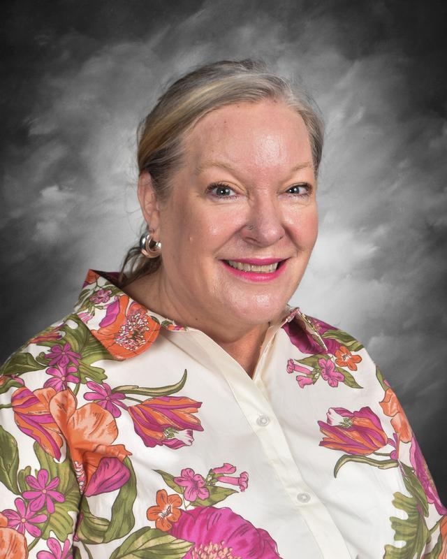 Smiling woman with shoulder-length hair, wearing a colorful floral shirt, against a gray background.