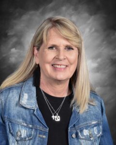 A smiling woman with blonde hair wearing a denim jacket and layered necklaces against a gray background.
