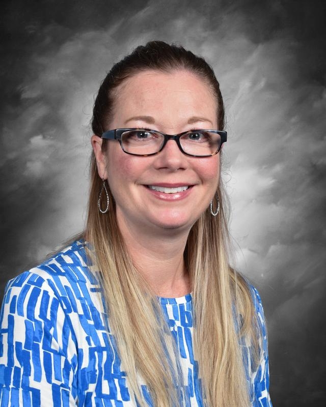 Smiling woman with long, straight hair wearing glasses and a blue patterned blouse against a gray background.