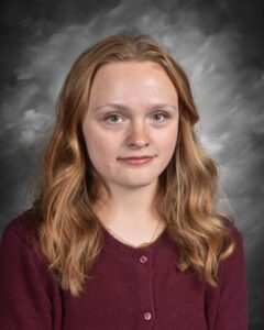 A young woman with wavy, light brown hair wearing a maroon cardigan, posed against a gray backdrop.
