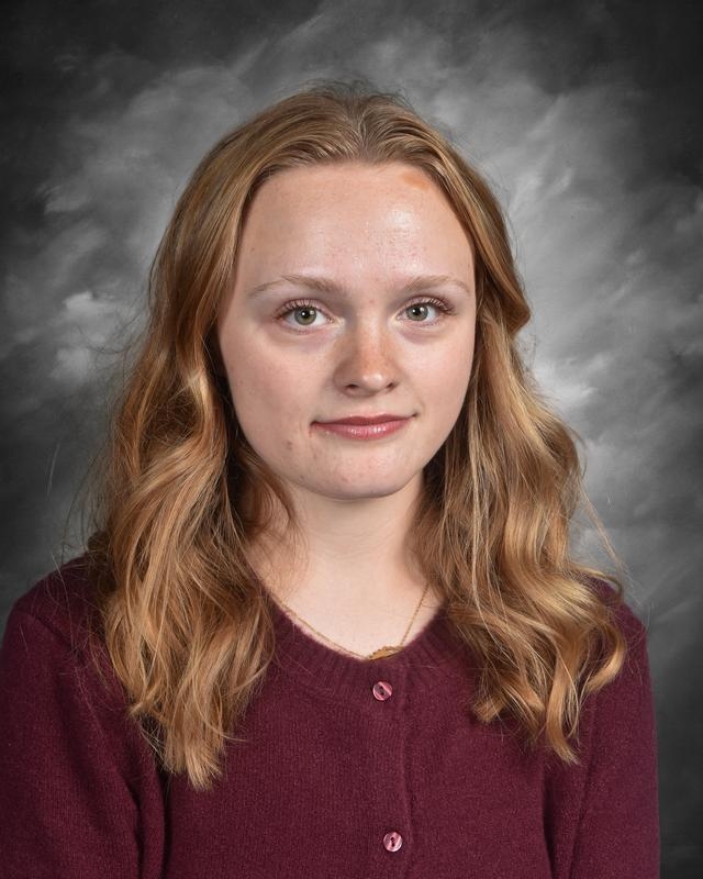 A young woman with wavy, light brown hair wearing a maroon cardigan, posed against a gray backdrop.