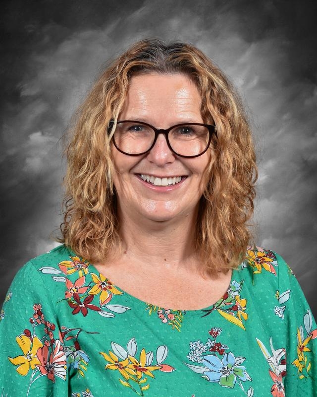 Smiling woman with curly hair, wearing glasses and a floral green blouse, against a gray background.