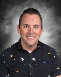 Smiling man with short hair, wearing a black shirt with planet and astronaut patterns, against a gray background.