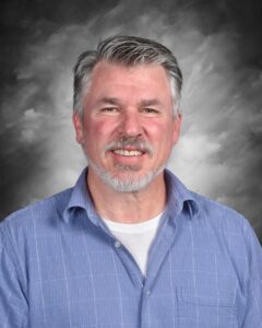 Smiling man with gray hair and beard, wearing a blue button-up shirt, against a gray background.