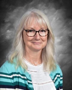 A woman with long, light hair and glasses smiles, wearing a teal and white striped cardigan against a gray background.