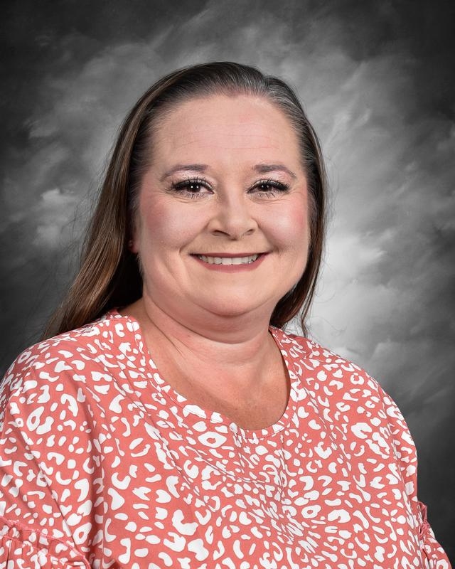 A smiling woman with long hair, wearing a pink patterned blouse, against a gray background.