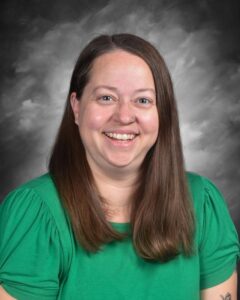 Smiling woman with long brown hair wearing a green shirt, against a gray background.