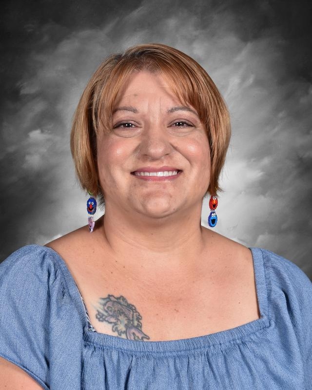 Smiling woman with short brown hair, wearing blue earrings and a blue top, posing against a gray background.