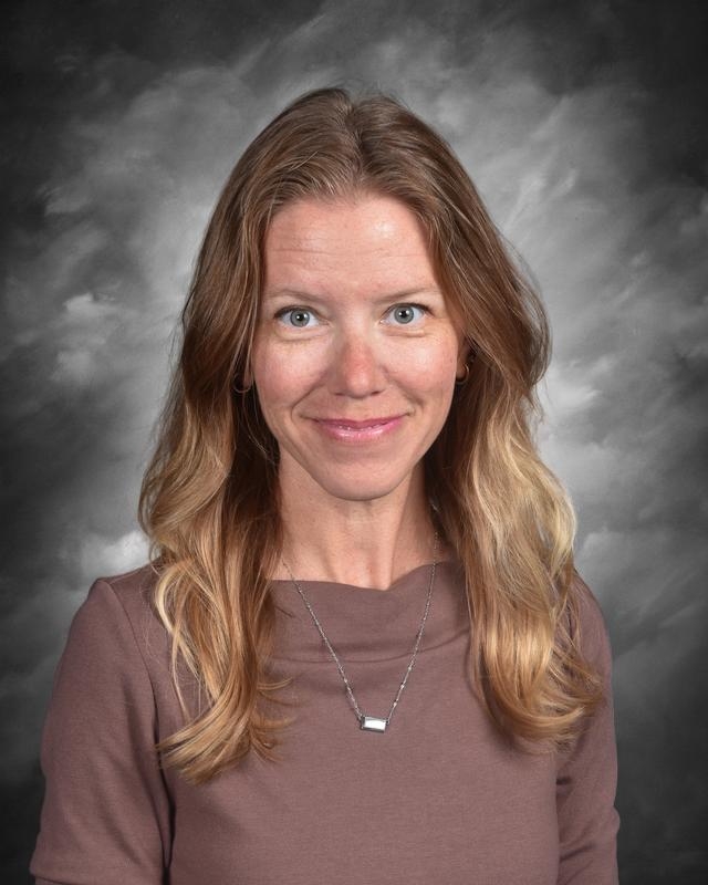 A woman with long, wavy hair smiles at the camera, wearing a brown top and a simple necklace, against a gray background.