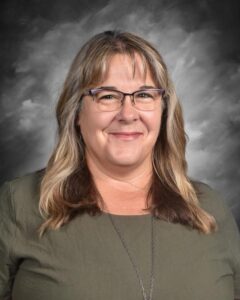 A smiling woman with long, wavy hair, wearing glasses and a green top, against a gray background.