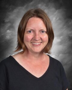 Una mujer con el pelo castaño hasta los hombros sonríe, vestida con una camiseta negra, sobre un fondo gris.