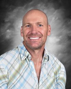 Smiling man with a shaved head and beard, wearing a plaid shirt, against a gray background.