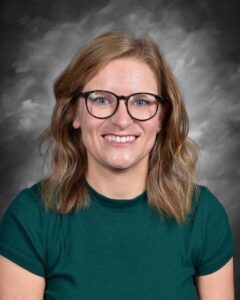 Smiling woman with wavy hair, wearing glasses and a green shirt, against a gray background.