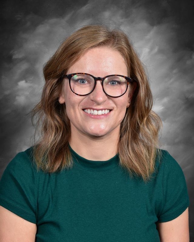 Smiling woman with wavy hair, wearing glasses and a green shirt, against a gray background.