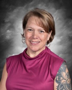 Smiling woman with short hair wearing a purple top, showcasing a detailed tattoo on her arm, against a gray backdrop.
