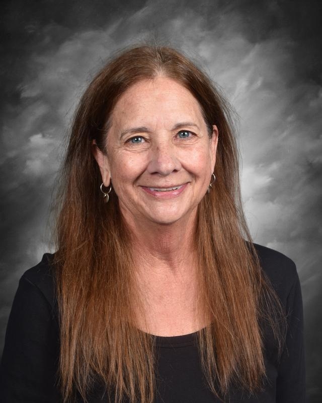 A smiling woman with long brown hair and earrings, wearing a black top, against a gray background.