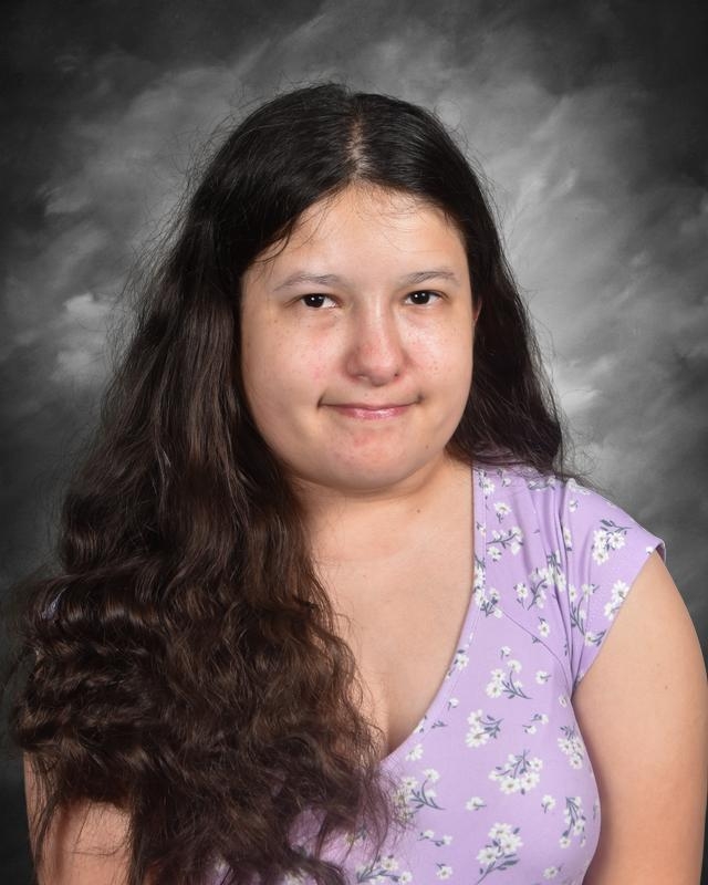 A young woman with long, wavy hair smiles gently, wearing a purple floral top against a gray backdrop.