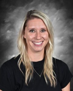 A smiling woman with long, blonde hair wearing a black shirt, set against a gray, cloudy background.