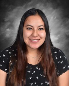 A smiling young woman with long dark hair, wearing a black floral blouse, against a gray background.