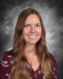 Smiling woman with long brown hair, wearing a floral blouse, against a soft gray background.