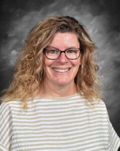 Smiling woman with curly hair, wearing glasses and a striped shirt, against a gray background.