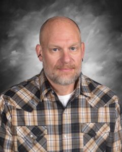 A man with a beard and short hair, wearing a plaid shirt, poses against a dark, cloudy background.