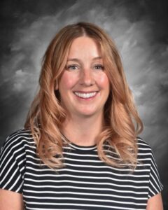 Smiling woman with wavy, shoulder-length hair wearing a striped shirt, set against a gray background.