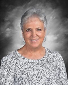 Smiling older woman with short gray hair, wearing a dotted blouse, against a soft gray background.
