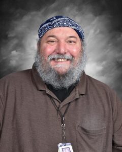 Smiling man with a beard and bandana, wearing a brown shirt and ID badge, set against a gray background.