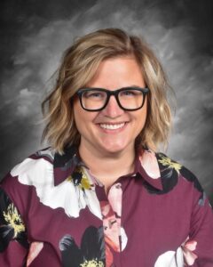 Smiling person with short, wavy hair wearing glasses and a floral shirt, set against a gray background.