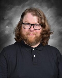 A man with long, curly hair and a beard smiles, wearing glasses and a black polo shirt against a gray backdrop.