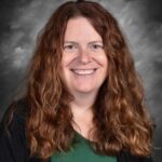 Smiling woman with long, curly brown hair, wearing a green shirt and black cardigan, against a gray backdrop.
