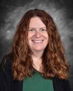 Smiling woman with long, curly brown hair, wearing a green shirt and black cardigan, against a gray backdrop.