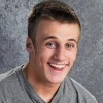 Smiling young man with short hair, wearing a gray shirt, set against a textured gray background.