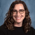 Smiling woman with curly hair wearing glasses, in a black shirt, against a blue backdrop.