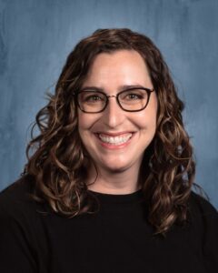 Smiling woman with curly hair wearing glasses, in a black shirt, against a blue backdrop.