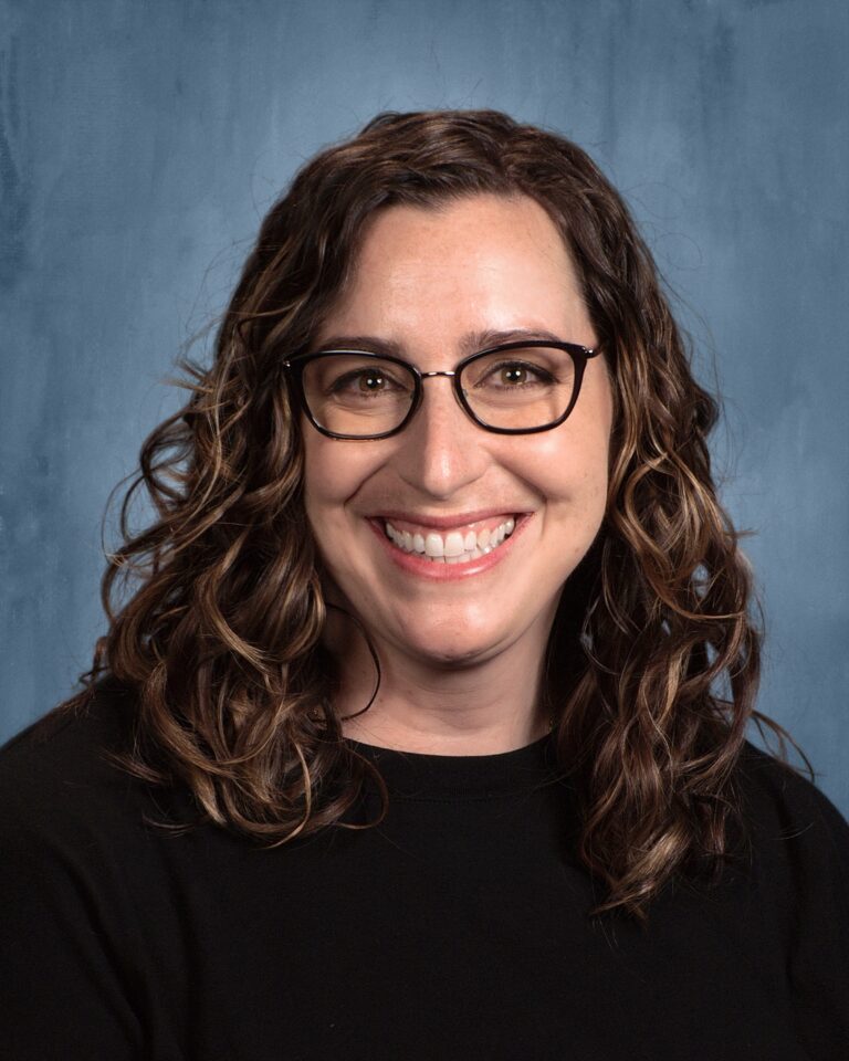 Smiling woman with curly hair wearing glasses, in a black shirt, against a blue backdrop.
