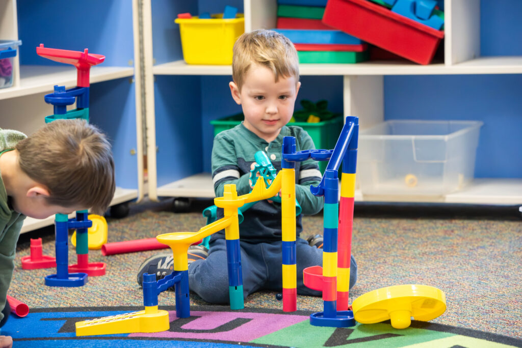 Two children play on a colorful rug with a marble run toy, one focusing on building while the other observes nearby.