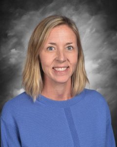 A smiling woman with shoulder-length blonde hair, wearing a blue sweater, poses against a gray backdrop.